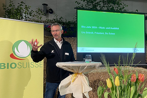 Un conférencier devant une présentation Power Point et un rollup, au premier plan une décoration florale.