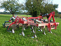 Machine à pattes d'oie servant à nettoyer les chaumes d'une parcelle après les moissons, elle est rouge et posée dans un pâturage.