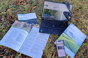 Ein Laptop mit einem Kopfhörer, Broschüren und einem Flyer steht auf einer Wiese. 