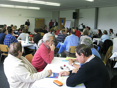 Photo-portrait de Christien Bieri.
Salle avec personnes en train de faire un travail en groupe, les personnes sont par quatre autour des tables.
Photo-portrait de Vitus Schafer