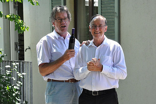 Deux hommes sourient à la caméra, l'homme de gauche tient une bouteille de vin.