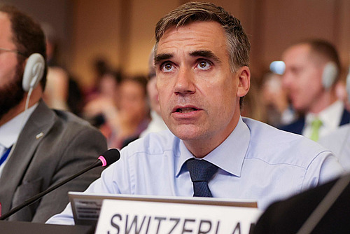 Un homme dans une salle avec un micro de table et un panneau indiquant «Switzerland».