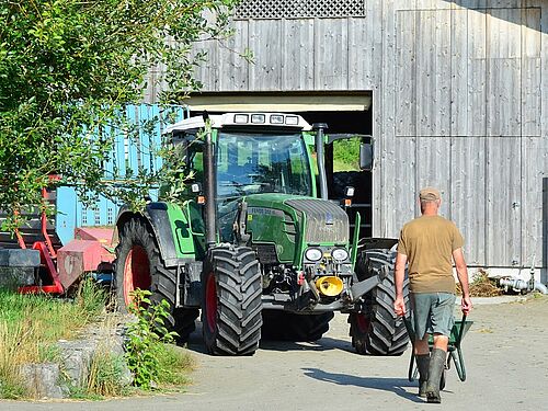 Un fermier marche avec une brouette et le dos tourné à la caméra vers le tracteur et la grange