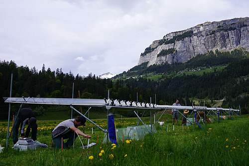 Devant un sommet alpin, des abris contre la pluie sont dressés dans un pré ; en dessous, des personnes sont à genoux et travaillent sur le sol. 