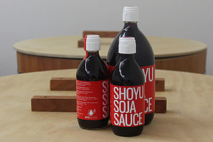 Sur une table se trouvent trois bouteilles foncées de tailles différentes, ornées d'une étiquette rouge portant l'inscription « Shoyu Soja Sauce ».  