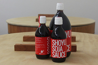 Sur une table se trouvent trois bouteilles foncées de tailles différentes, ornées d'une étiquette rouge portant l'inscription « Shoyu Soja Sauce ».  
