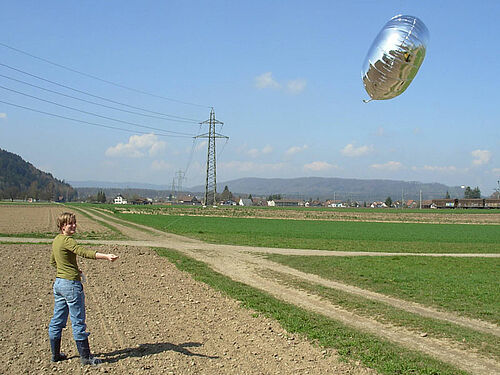 Frau hält silbrig-grauen Ballon in der Schwebe über einem frisch gesäten Maisacker