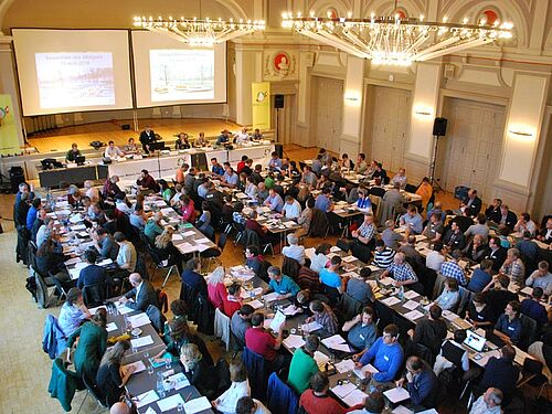 [Translate to Französisch:] Die Delegierten an der Arbeit im Saal mit zwei Kronleuchten und zwei bebeamten Leinwänden