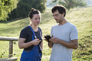 Une jeune femme et un jeune homme regardent ensemble le téléphone portable que le jeune homme tient dans ses mains. Ils se trouvent à l'extérieur, dans un champ.