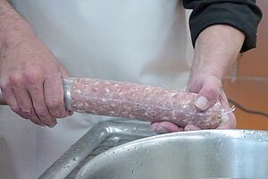 Zwei Hände halten an einer Wurstmaschine eine Salami, die mit Inhalt gefüllt wird.