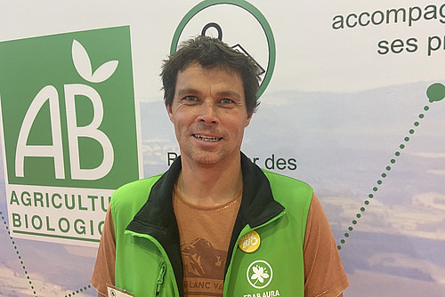 Ein Mann vor einer Reklamewand. Er trägt ein hellgrünes Gilet. 