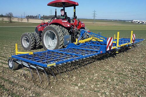 Herse-étrille bleue et jaune derrière tracteur rouge