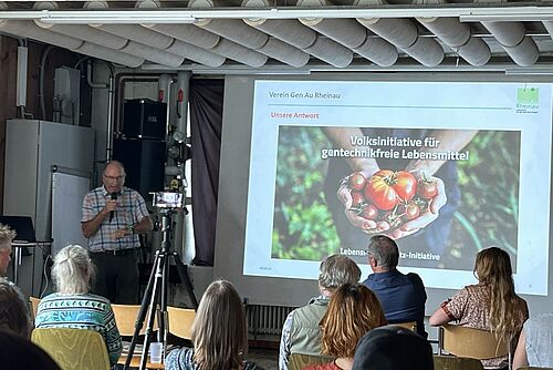 L'orateur s'exprime devant un public assis ; en arrière-plan, un transparent de présentation avec le texte Initiative populaire pour des aliments produits sans manipulations génétiques.