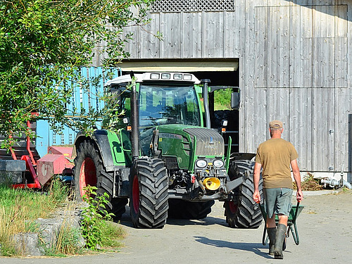 Un fermier marche avec une brouette et le dos tourné à la caméra vers le tracteur et la grange