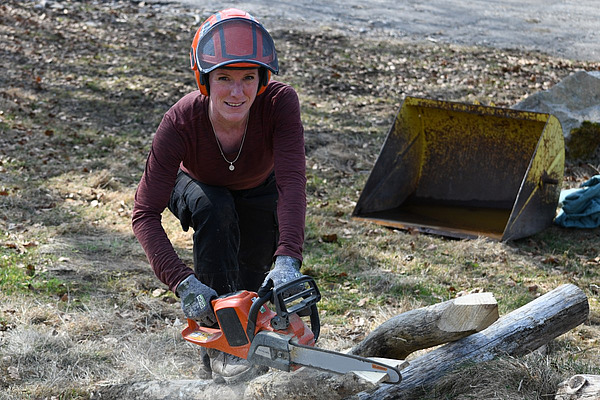 Une femme portant un casque tient une tronçonneuse à la main et taille le bout d'un poteau de clôture.