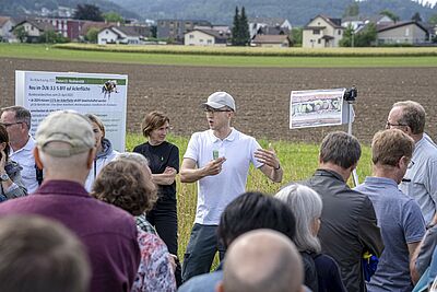 Un orateur fait des gestes au bord d'un terrain devant un groupe d'auditeurs.