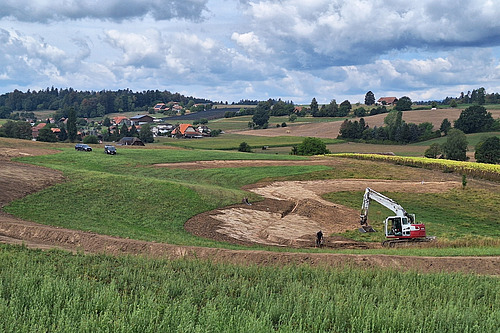 On aperçoit une vallée à caractère agricole. Au premier plan, des travaux de terrassement à grande échelle sont effectués à l'aide d'une pelleteuse.