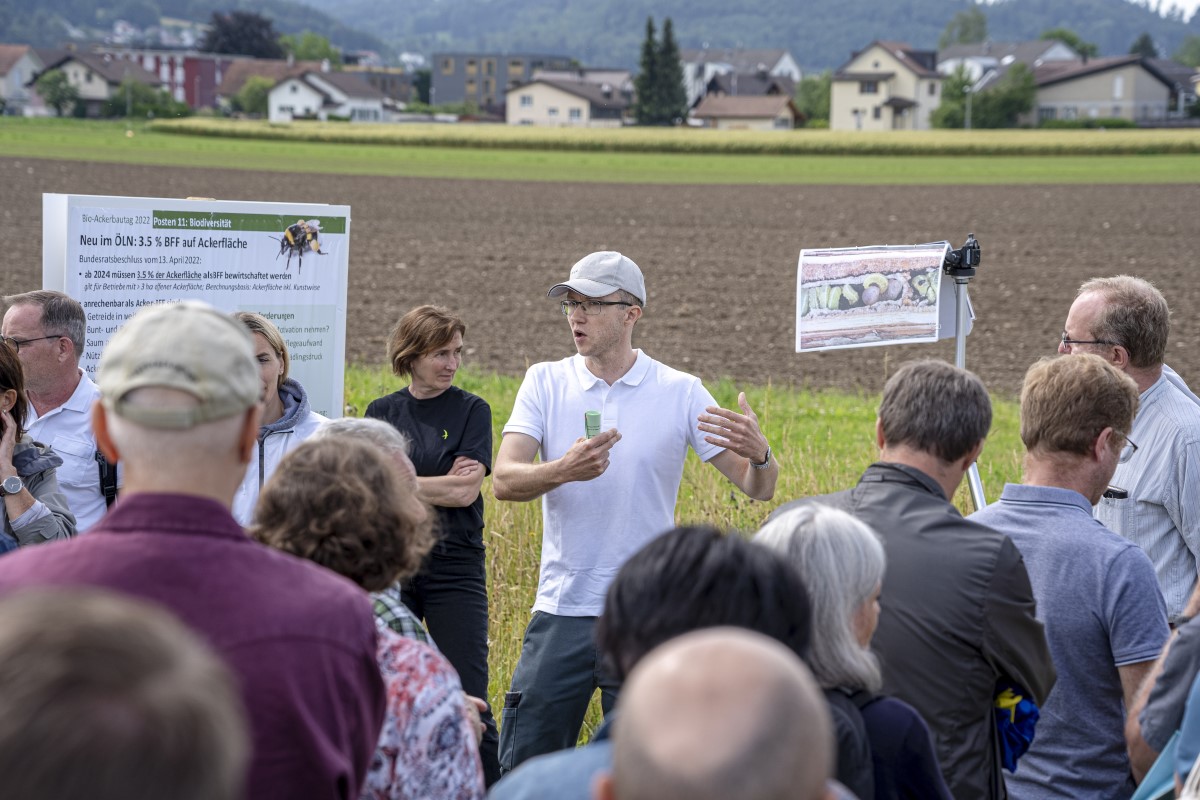 Un orateur fait des gestes au bord d'un terrain devant un groupe d'auditeurs.